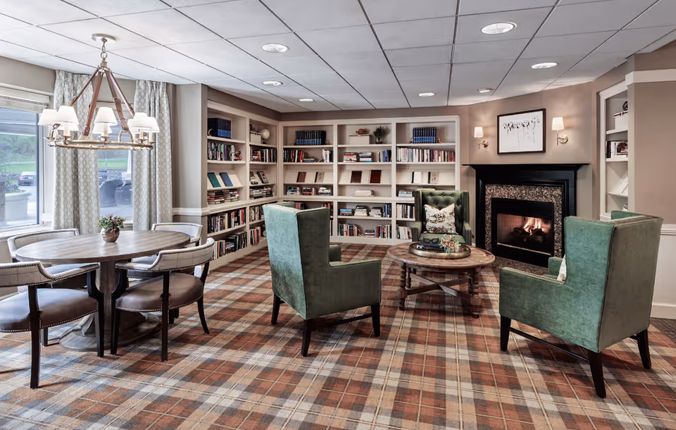 A cozy common area with a plaid carpet, three green upholstered armchairs around a wooden coffee table, a fireplace with a fire burning, and built-in bookshelves filled with books. To the left, there is a round wooden table with six cushioned chairs and a chandelier above it. Large windows with curtains allow natural light into the room.