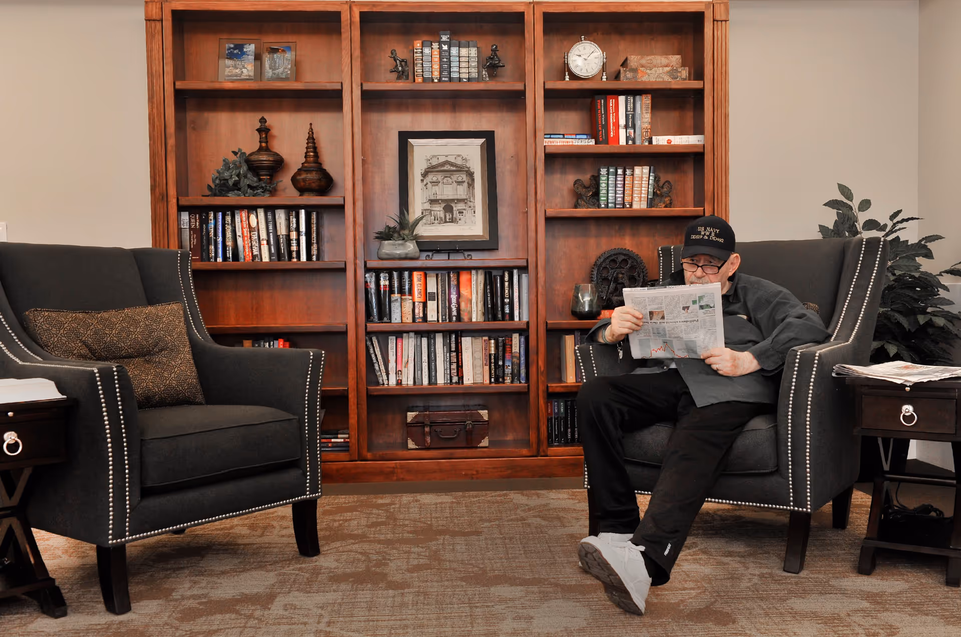 An elderly man wearing a black cap and glasses is sitting in a dark gray armchair reading a newspaper. Another matching armchair with a decorative pillow is positioned to the left. Behind them is a large wooden bookshelf filled with books, decorative items, and a framed picture. A small side table with a drawer and a plant are also visible in the room.