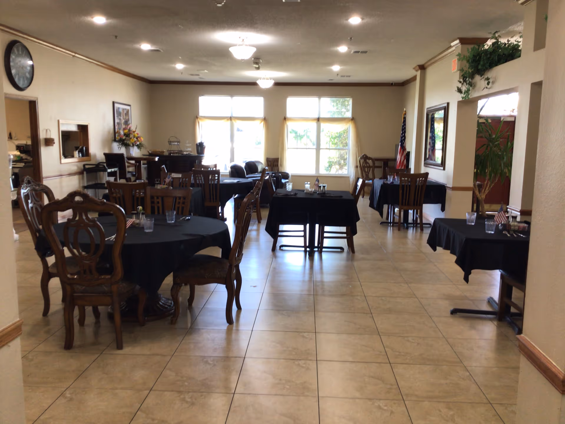 Senior living dining room with round tables, black tablecloths, wooden chairs, windows, and an American flag.
