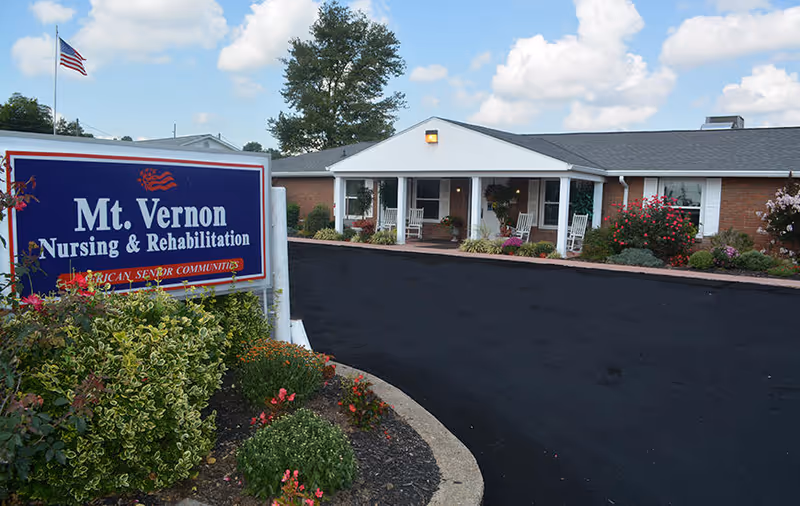 Front entrance of Mt. Vernon Nursing & Rehabilitation with a large sign, landscaped flowerbeds, and rocking chairs on the covered porch.