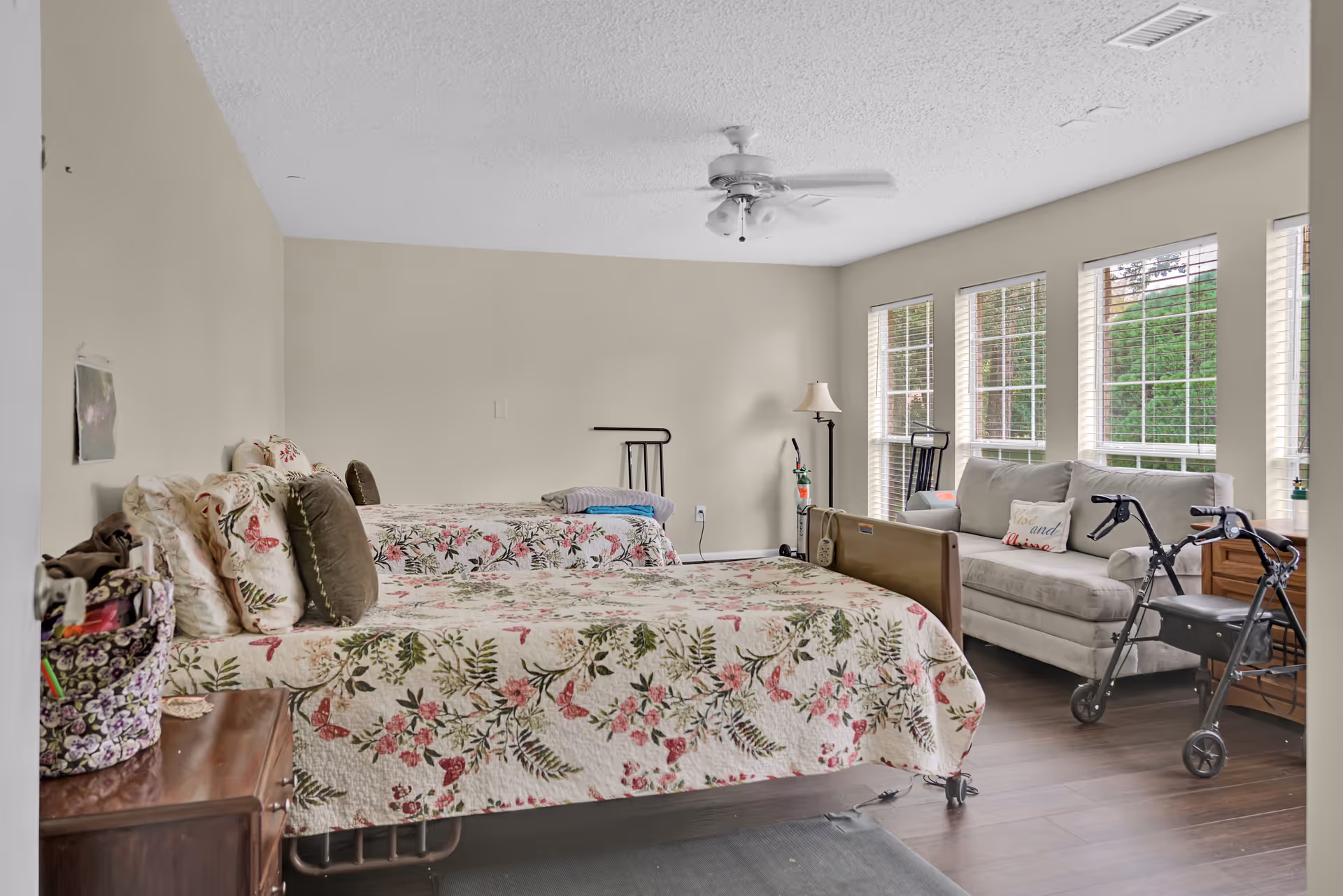 Bright shared bedroom with two floral-patterned twin beds, a sofa by large windows, and mobility aids like a walker and cane.
