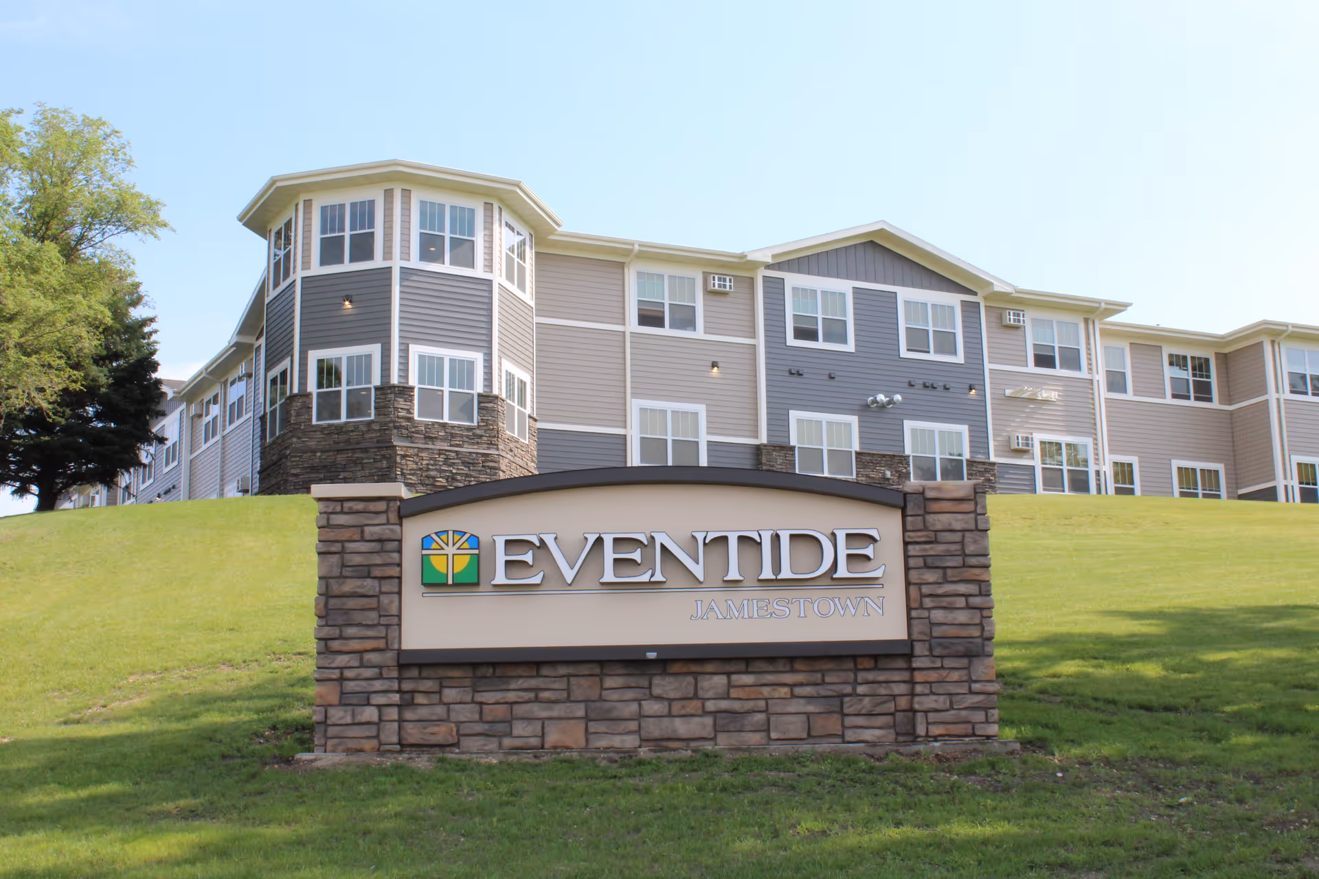 A stone sign reading 'EVENTIDE JAMESTOWN' on a grassy hill in front of a multi-story senior living building.