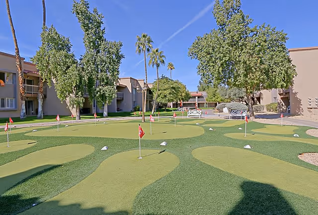 Outdoor putting green area with multiple small flags and holes, surrounded by trees and beige residential buildings under a clear blue sky.