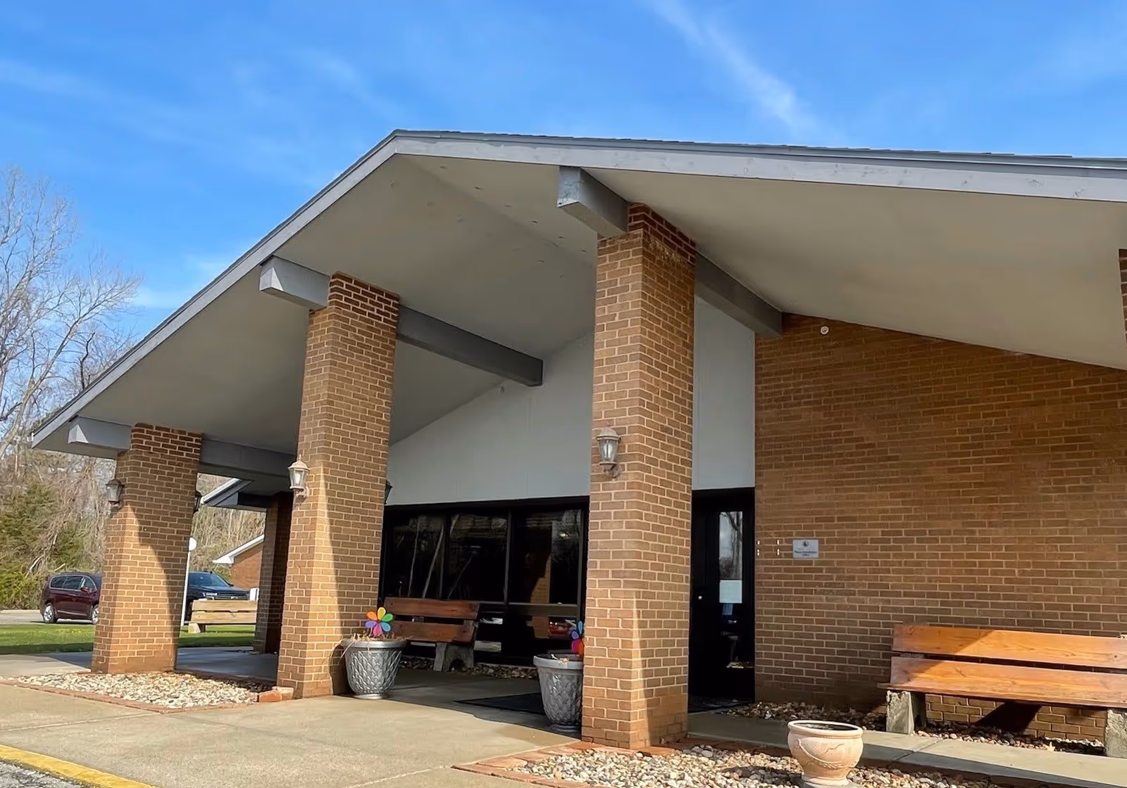 Exterior view of Carriage Square Rehab & Healthcare Center showing a brick building with a covered entrance supported by brick pillars. There are benches and large planters with colorful pinwheels near the entrance. The sky is clear and blue.