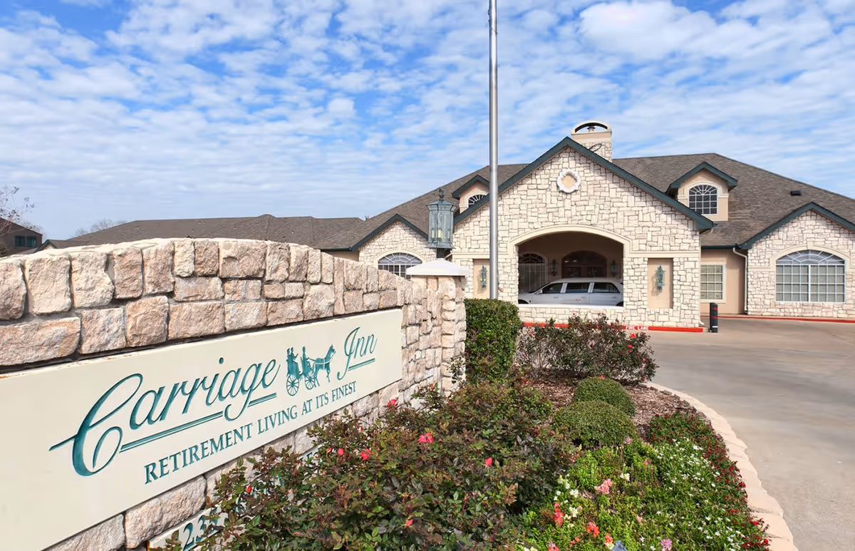 Exterior view of Carriage Inn at Bryan, a retirement living facility with a stone facade building, a driveway, and a landscaped garden with flowers and bushes in front of a stone sign displaying the facility's name.