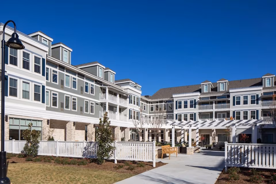 Exterior view of a multi-story senior living facility building with gray and white siding, multiple windows, balconies, a white pergola, benches, and a fenced landscaped courtyard under a clear blue sky.