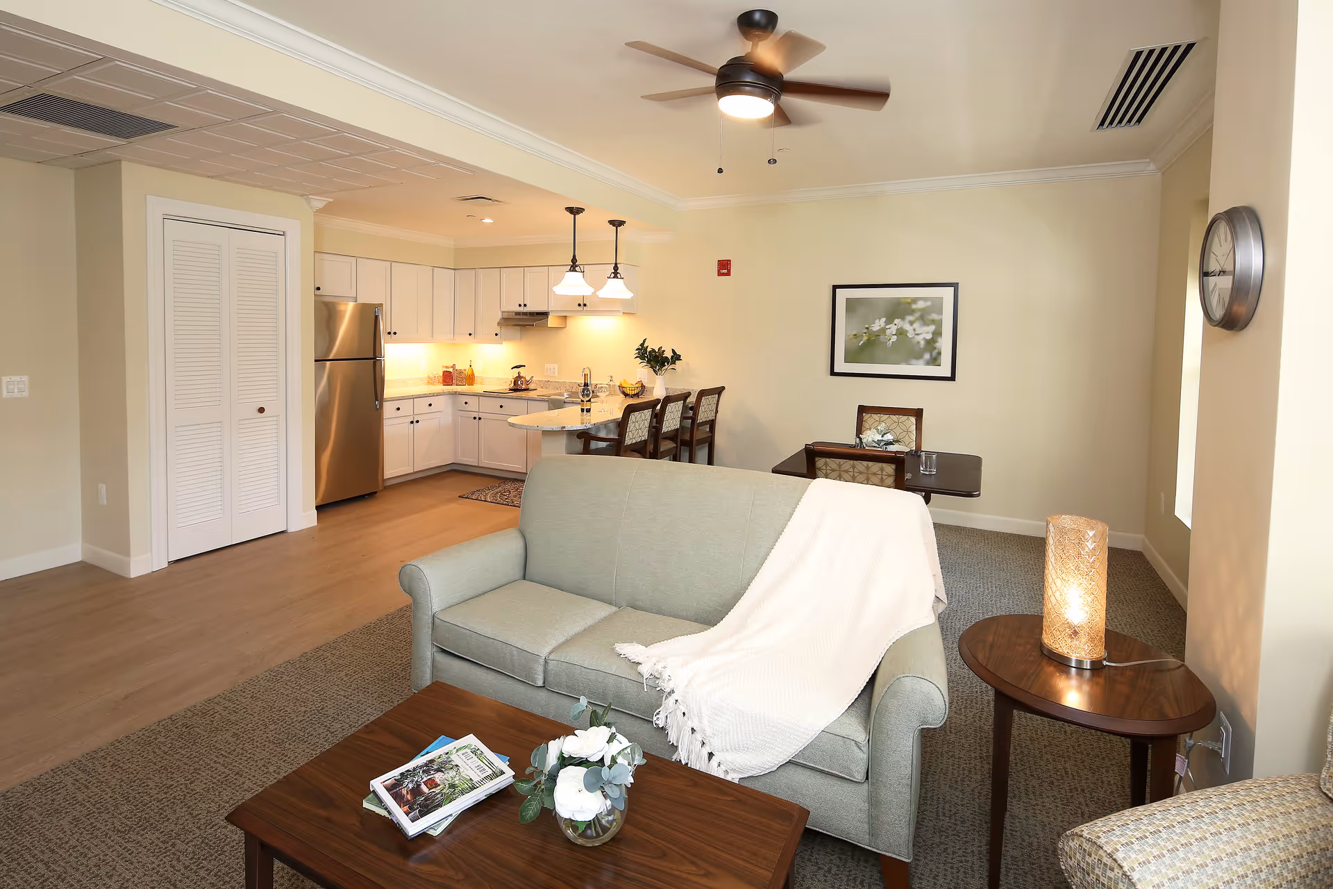A bright and cozy senior living apartment interior featuring a light green sofa with a white throw blanket, a wooden coffee table with magazines and a flower vase, a small dining table with two chairs, and a kitchen area with white cabinets, stainless steel refrigerator, and a breakfast bar with three chairs. The room has beige walls, a ceiling fan with light, a wall clock, and a decorative lamp on a side table.