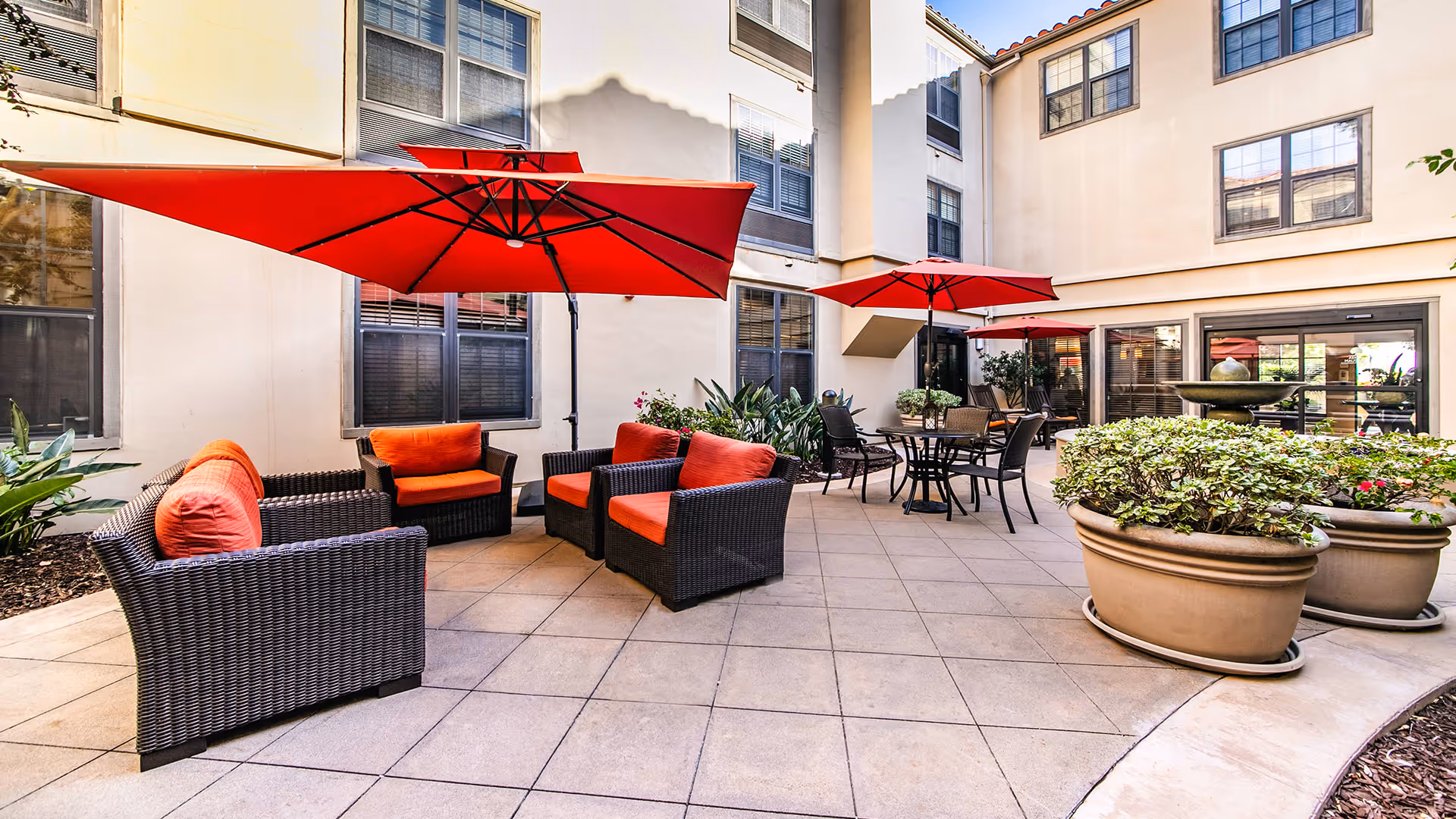 Outdoor courtyard area with wicker seating featuring orange cushions, red umbrellas providing shade, round tables with chairs, large potted plants, and surrounding building windows.