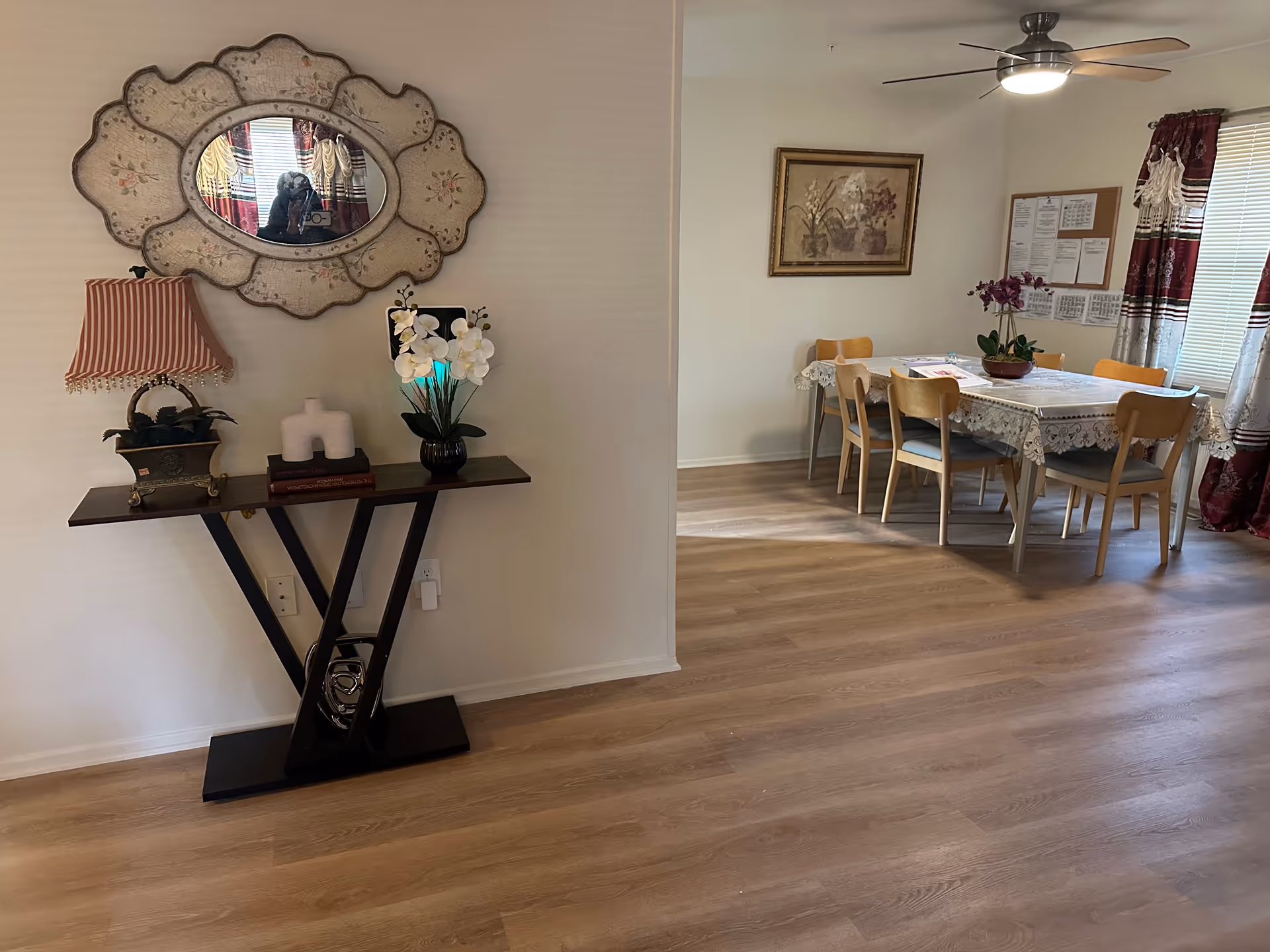 Interior view of a senior living facility showing a decorative console table with a lamp, books, and flowers beneath an ornate wall mirror on the left. On the right, there is a dining area with a table covered by a lace tablecloth, six wooden chairs, a ceiling fan with light, a framed floral painting on the wall, and windows with red and white curtains.
