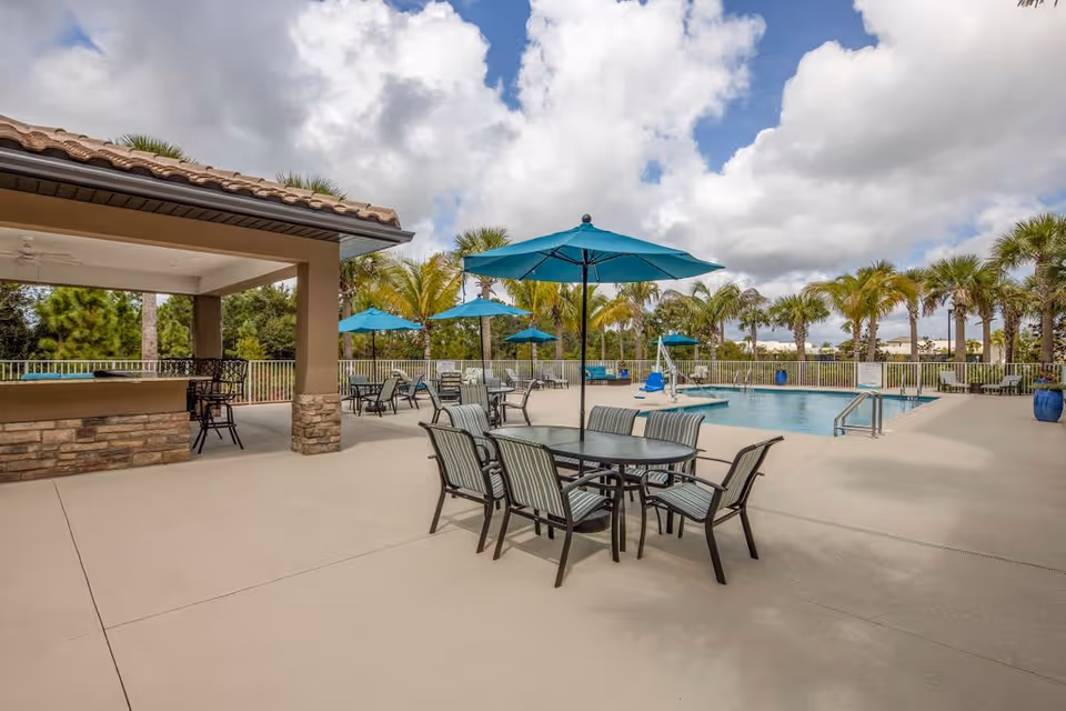 Outdoor pool area with several tables and chairs under blue umbrellas, a covered seating area with bar stools, palm trees in the background, and a partly cloudy sky.