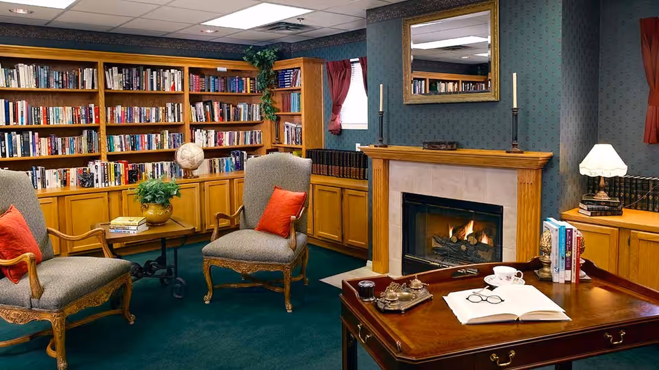 A cozy library room with wooden bookshelves filled with books, two upholstered armchairs with red cushions, a wooden coffee table with an open book and reading glasses, a fireplace with a fire burning, and a lamp on a side table. The room has green carpet and patterned wallpaper with a window covered by red curtains.