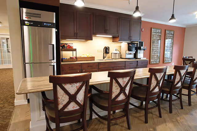 Communal dining area with a long wooden counter and six upholstered chairs facing a kitchen wall with dark cabinets, a stainless refrigerator and a coffee station.