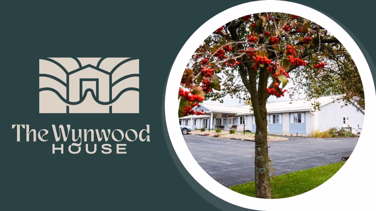 Exterior view of a single-story residential building with light blue siding and a white roof, partially obscured by a tree with red berries in the foreground. The building is surrounded by a paved driveway and some landscaping.