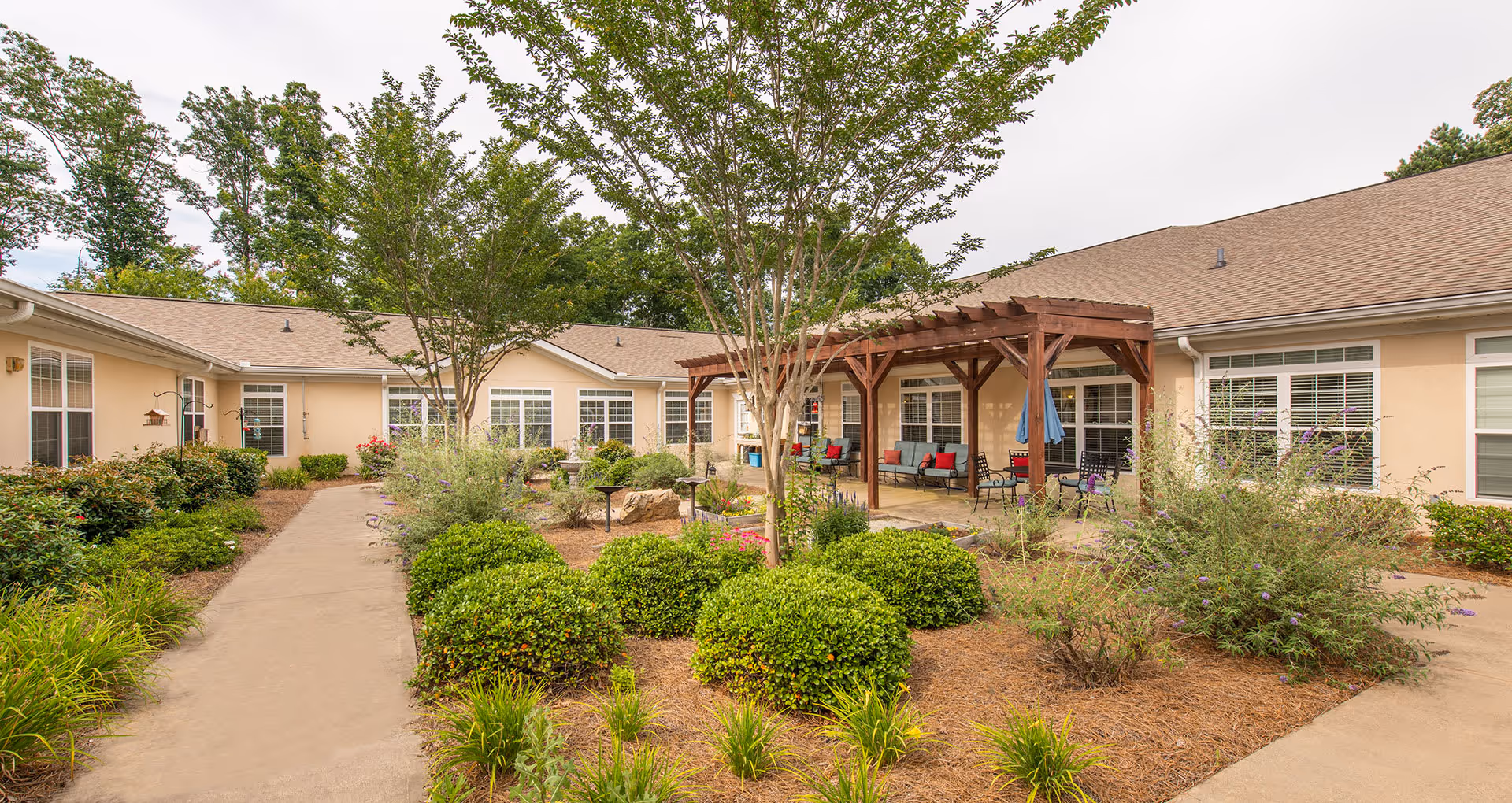 A landscaped outdoor courtyard at Benton House of Sugar Hill featuring a paved walkway, neatly trimmed bushes, flowering plants, and trees. There is a wooden pergola with seating underneath, and the beige building with multiple windows surrounds the courtyard.