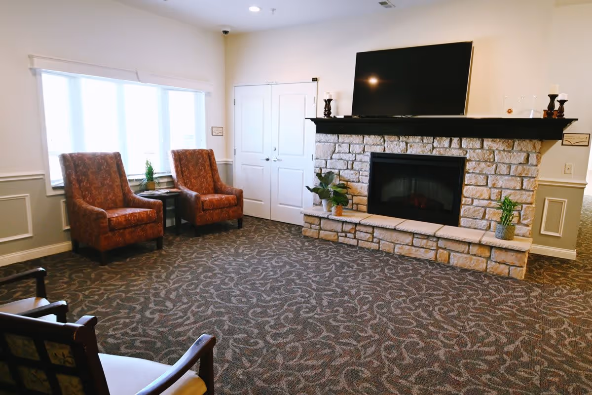 A cozy living room area with a stone fireplace and a large flat-screen TV mounted above it. Two patterned armchairs are placed near a window with white blinds, and there are small plants on the fireplace hearth and a side table between the chairs. The room has patterned carpet and light-colored walls with white trim.