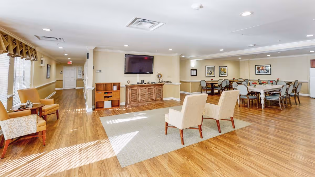 A spacious senior living facility common area with wood flooring, beige walls, and recessed lighting. The room features two beige armchairs on a light area rug, a wooden cabinet with a flat-screen TV mounted above it, and several dining tables with chairs arranged around the room. Large windows with curtains allow natural light to enter, and framed artwork decorates the walls.