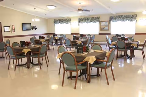 A spacious dining room with multiple round tables and green chairs in a senior living facility.