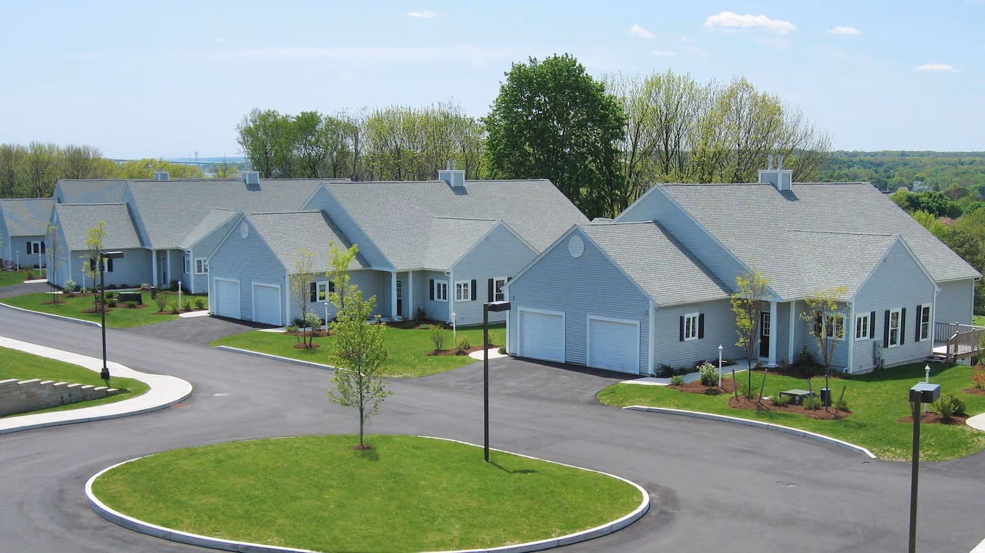 Aerial view of single-story light-gray residential buildings with attached garages, circular driveway, lawns, and lamp posts.
