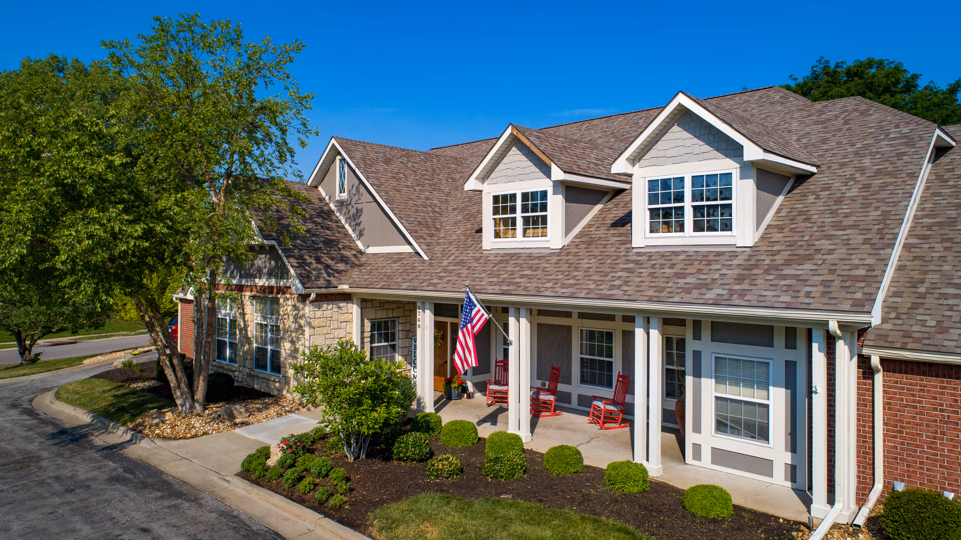 Front exterior of an assisted living building with a covered porch, red rocking chairs, an American flag, and landscaped shrubs.