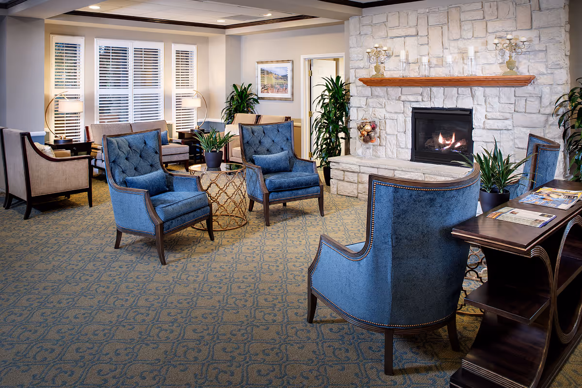 A cozy senior living facility lounge area with blue upholstered armchairs arranged around a small round table. The room features a stone fireplace with a wooden mantle, decorative plants, and a carpet with a subtle pattern. Large windows with white blinds allow natural light to fill the space, and there are additional seating areas with beige chairs and side tables with lamps.