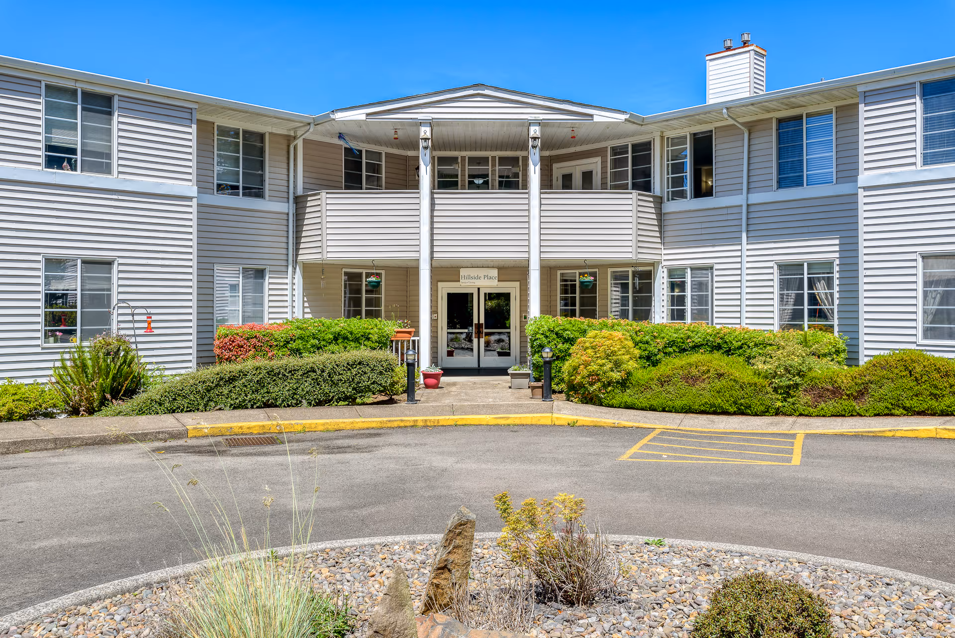 Exterior front view of Hillside Place Assisted Living & Memory Care building with white siding, multiple windows, a covered entrance with double glass doors, surrounded by green bushes and landscaping under a clear blue sky.