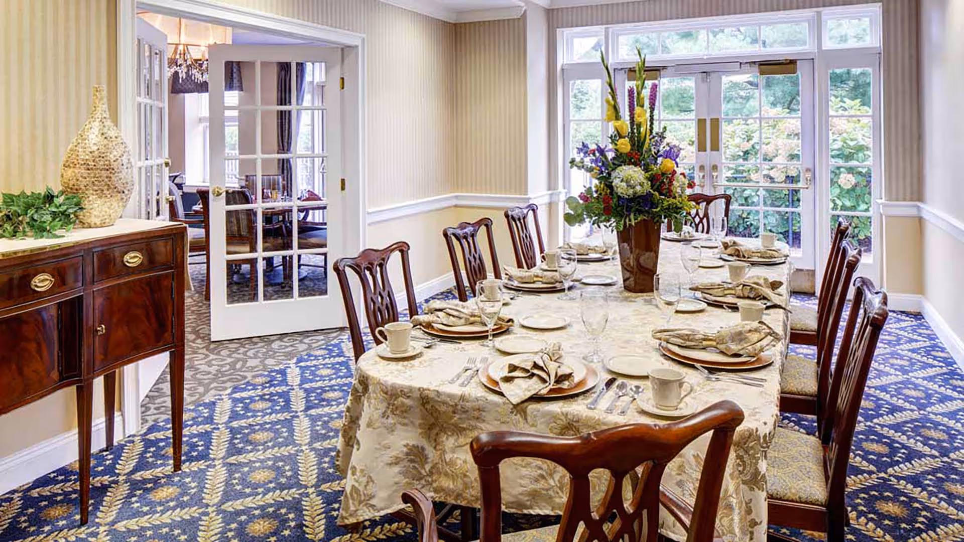 A formal dining room with a long table covered with a patterned tablecloth, set with plates, cups, napkins, and glassware. There is a large floral centerpiece on the table. The room has patterned carpet, cream-colored walls with white trim, and French doors leading to another room and to the outside garden area.