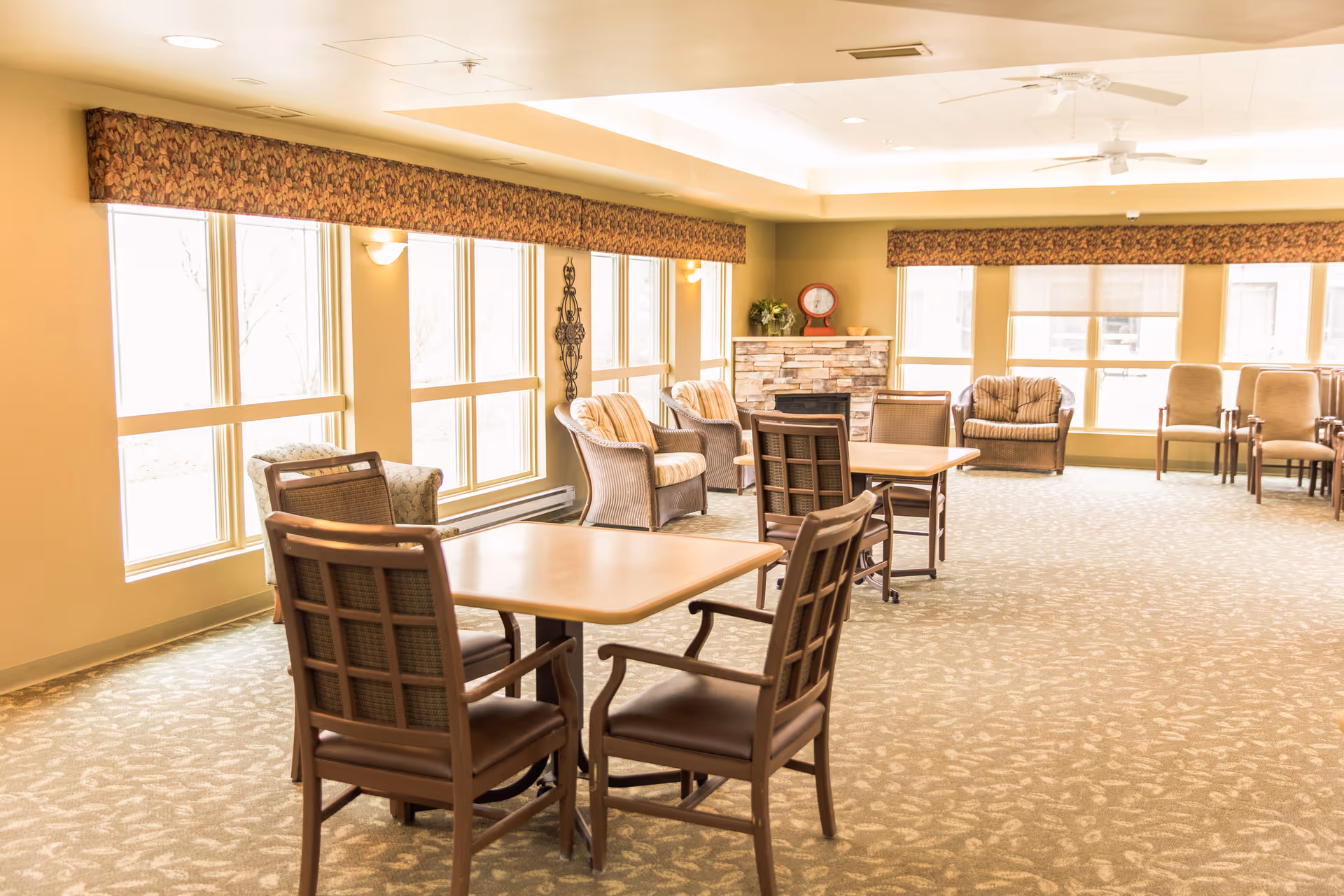 A bright and spacious common area in a senior living facility with multiple tables and chairs arranged for seating. Large windows allow natural light to fill the room. There are cushioned armchairs near a decorative stone fireplace, and the room features patterned carpet and ceiling fans.