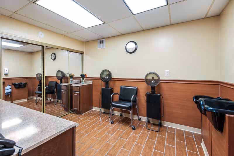 Interior room with wood-look tile flooring and beige walls featuring a wooden wainscot. The room contains two black salon chairs with attached hair dryers, a black wash basin, a cabinet with a small flower arrangement, and a large mirror with sliding doors reflecting the room.