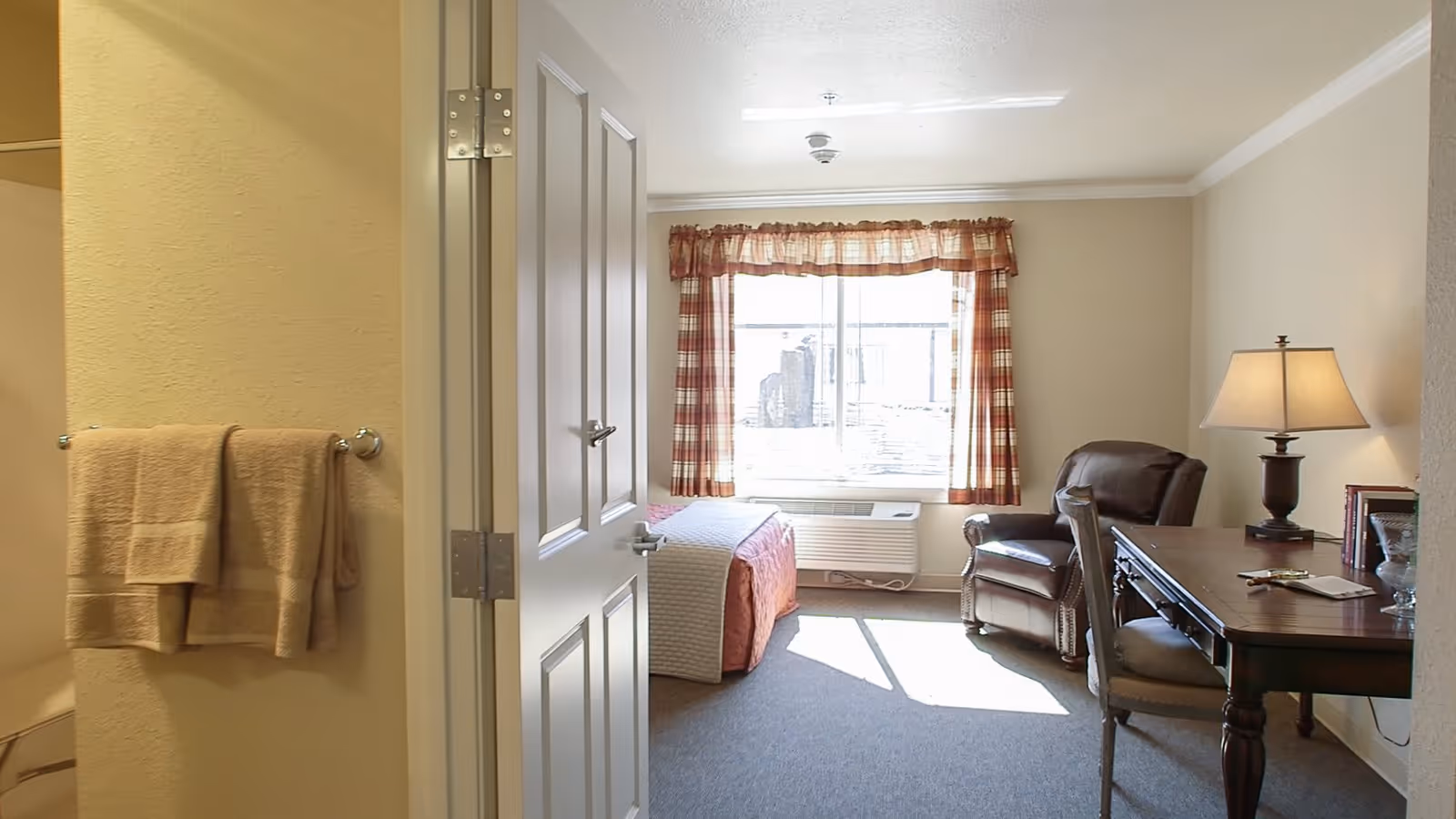 View of a senior living facility room with a bed near a window with plaid curtains, a brown leather armchair, a wooden desk with a lamp, and a chair. To the left, a bathroom entrance is visible with beige towels hanging on a towel rack.