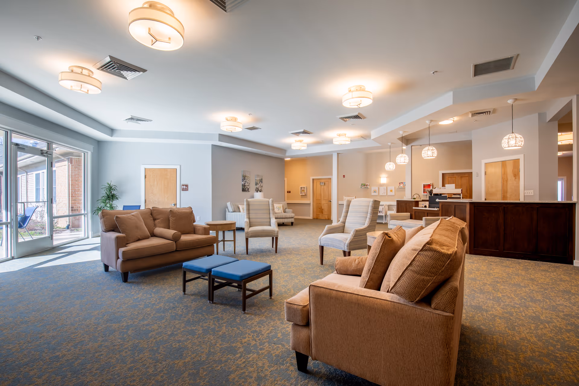Bright, open senior living facility common area with sofas, armchairs, a reception desk, and large windows.