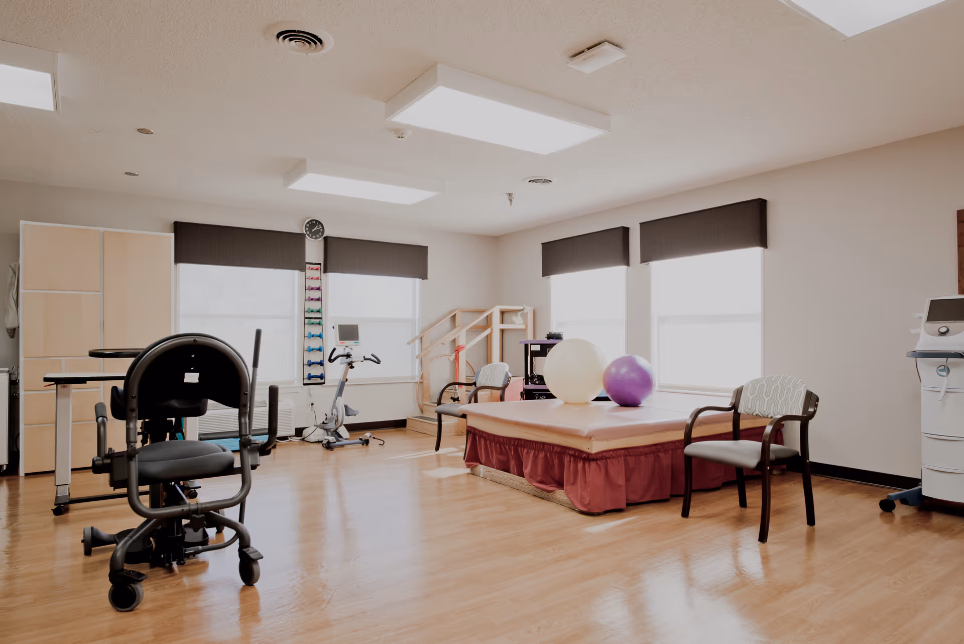 A bright physical therapy room with wooden flooring, exercise equipment including a stationary bike, therapy balls on a padded table, chairs, and a small set of stairs with handrails near the windows.
