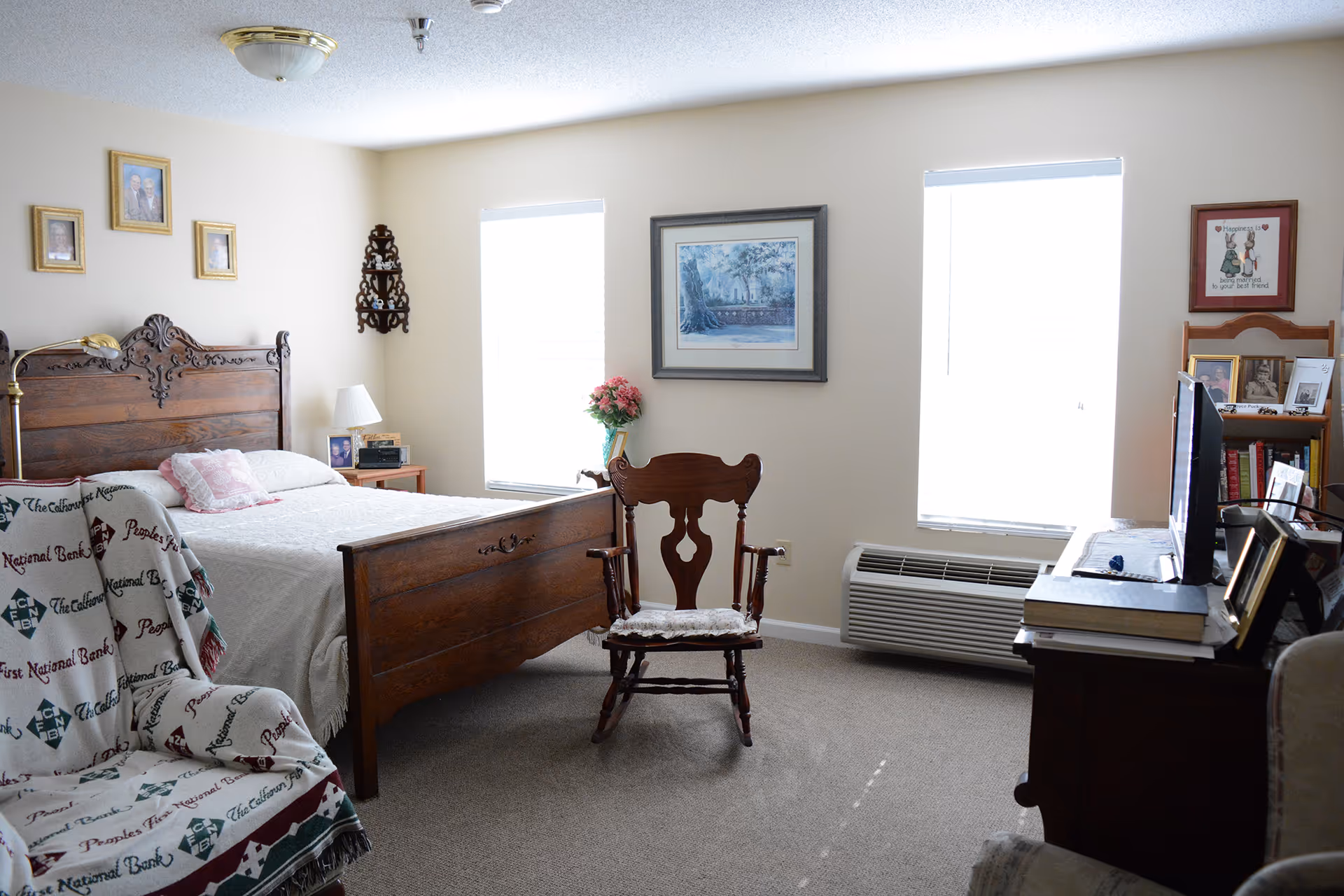 A cozy bedroom with a wooden bed frame, a rocking chair with a cushion, a small side table with a lamp, and a TV on a dresser. The room has two windows with blinds, framed pictures on the walls, and a patterned blanket draped over an armchair.