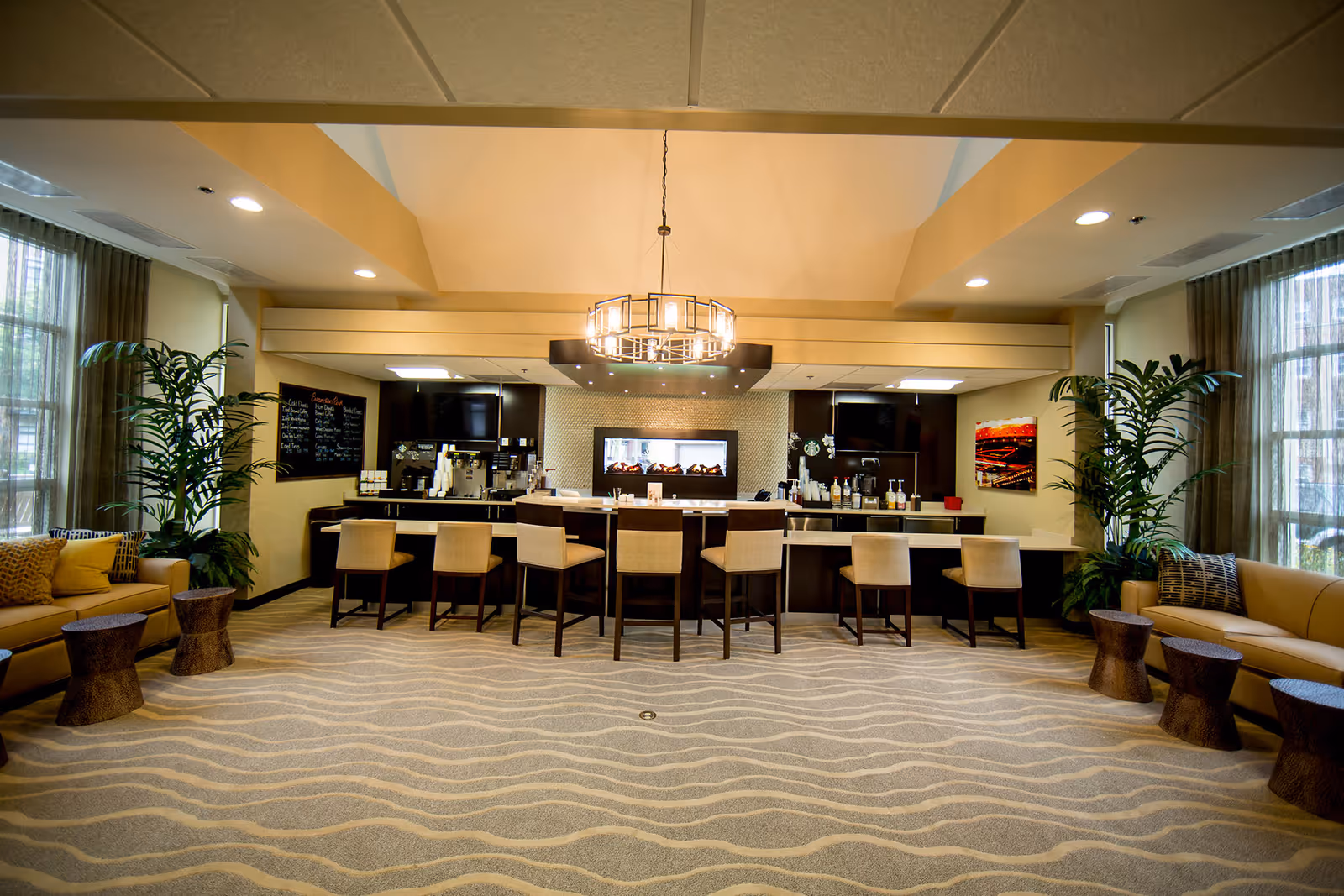 A spacious and well-lit lounge area with a modern coffee bar in the center, featuring seven bar stools. The room has large windows with sheer curtains on both sides, two beige sofas with cushions, and several small wooden side tables. There are tall green plants near the sofas and a contemporary chandelier hanging from the ceiling.