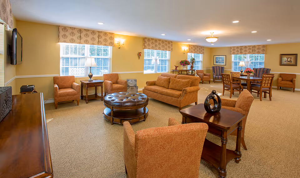 A spacious and well-lit living room area in a senior living facility with multiple comfortable upholstered chairs and sofas arranged around wooden tables. The room features large windows with patterned valances, beige walls, carpeted flooring, and decorative lamps and artwork. There is a round leather ottoman in the center and a flat-screen TV mounted on the wall.