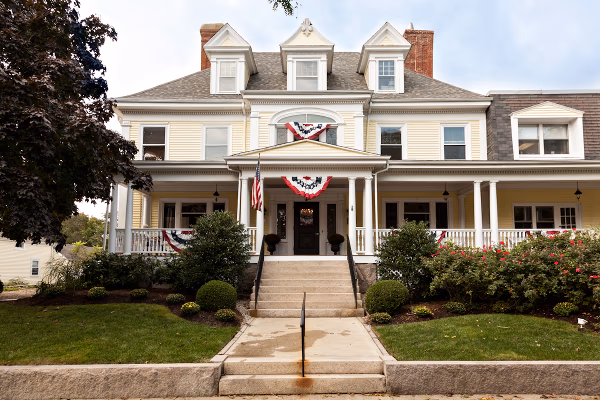 Front exterior view of a large, traditional-style yellow house with white trim and a porch decorated with red, white, and blue bunting. There are steps leading up to the porch, an American flag on a pole, and well-maintained bushes and flowers in the front yard.