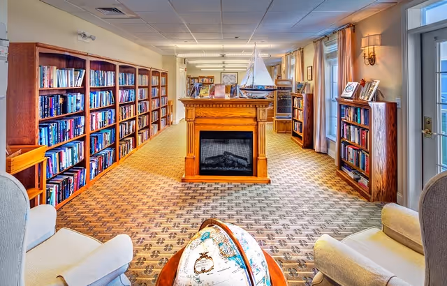 A bright and cozy library room with wooden bookshelves filled with books lining the walls. In the center is a wooden fireplace with a decorative sailboat on top. Two comfortable armchairs face each other near a globe on a wooden stand. Large windows with curtains allow natural light to fill the room.