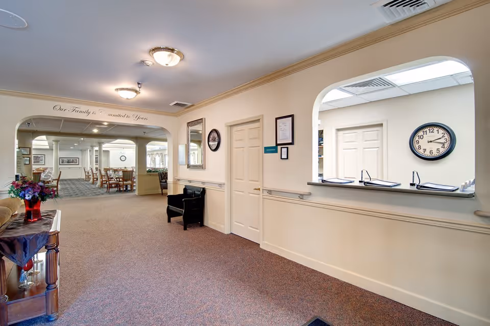 Interior view of a senior living facility hallway with beige walls and carpeted floor. There is a reception window with clipboards and a clock on the wall behind it. The hallway leads to a dining area with tables and chairs. A decorative table with a flower vase is on the left side, and a black chair is placed against the wall near two closed doors. Above the archway, a sign reads 'Our Family is Committed to Yours.'