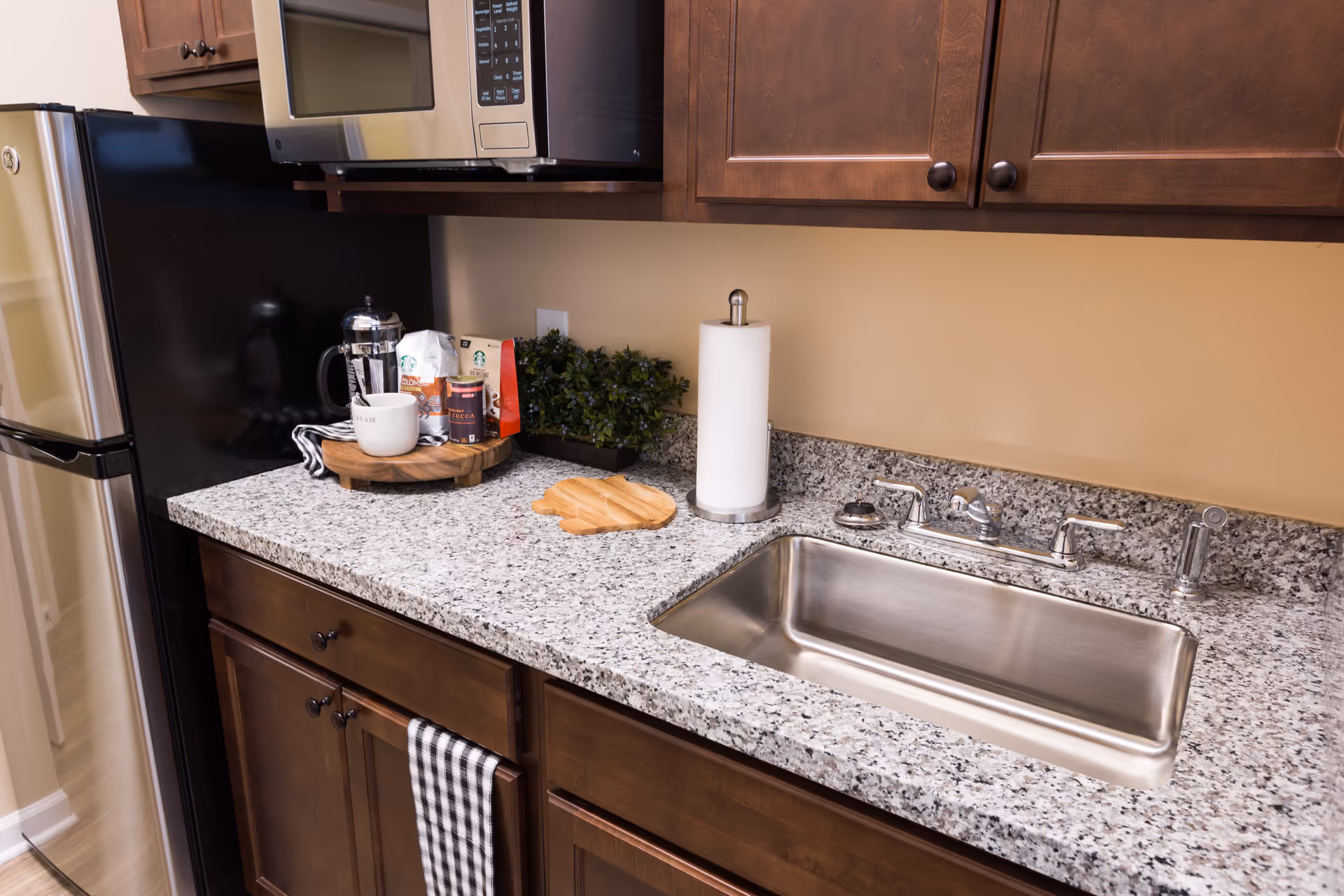 A kitchen countertop with a stainless steel sink, a paper towel holder, a small cutting board, a French press coffee maker, coffee bags, a white mug, and a small green plant. Above the countertop are wooden cabinets and a microwave. To the left is a stainless steel refrigerator.