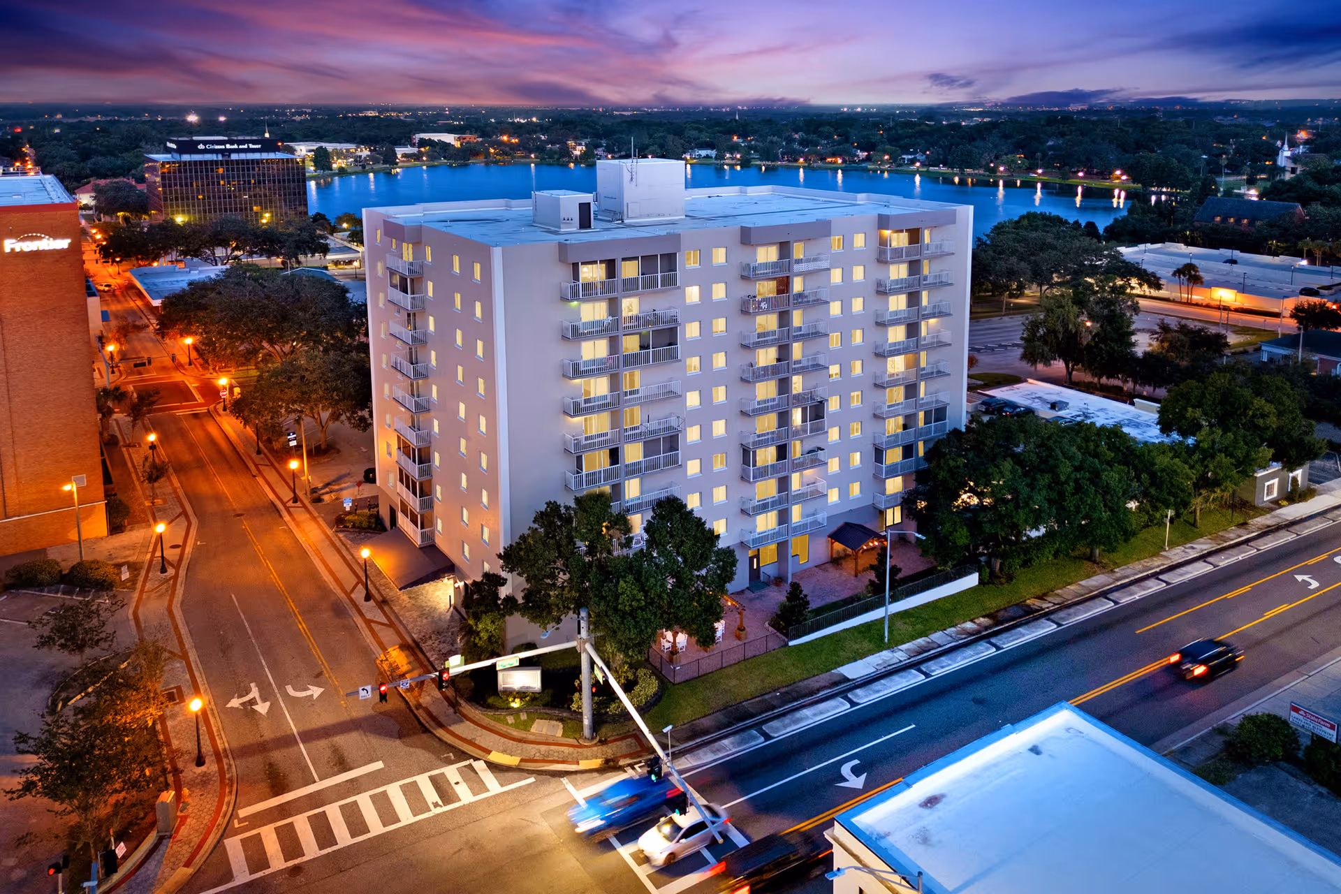 Aerial view of a multi-story residential building at dusk with lights on inside. The building is located at a street intersection with visible road markings and traffic lights. Trees and other buildings surround the area, and a body of water is visible in the background under a colorful evening sky.