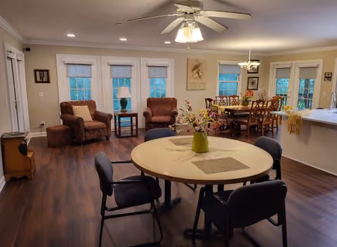 A spacious, well-lit room with wooden flooring featuring a round table with four black chairs in the foreground, two brown armchairs with a small side table and lamp near three windows in the background, and a dining table with several chairs further back. The room has beige walls, white trim, a ceiling fan with lights, and decorative artwork on the walls.