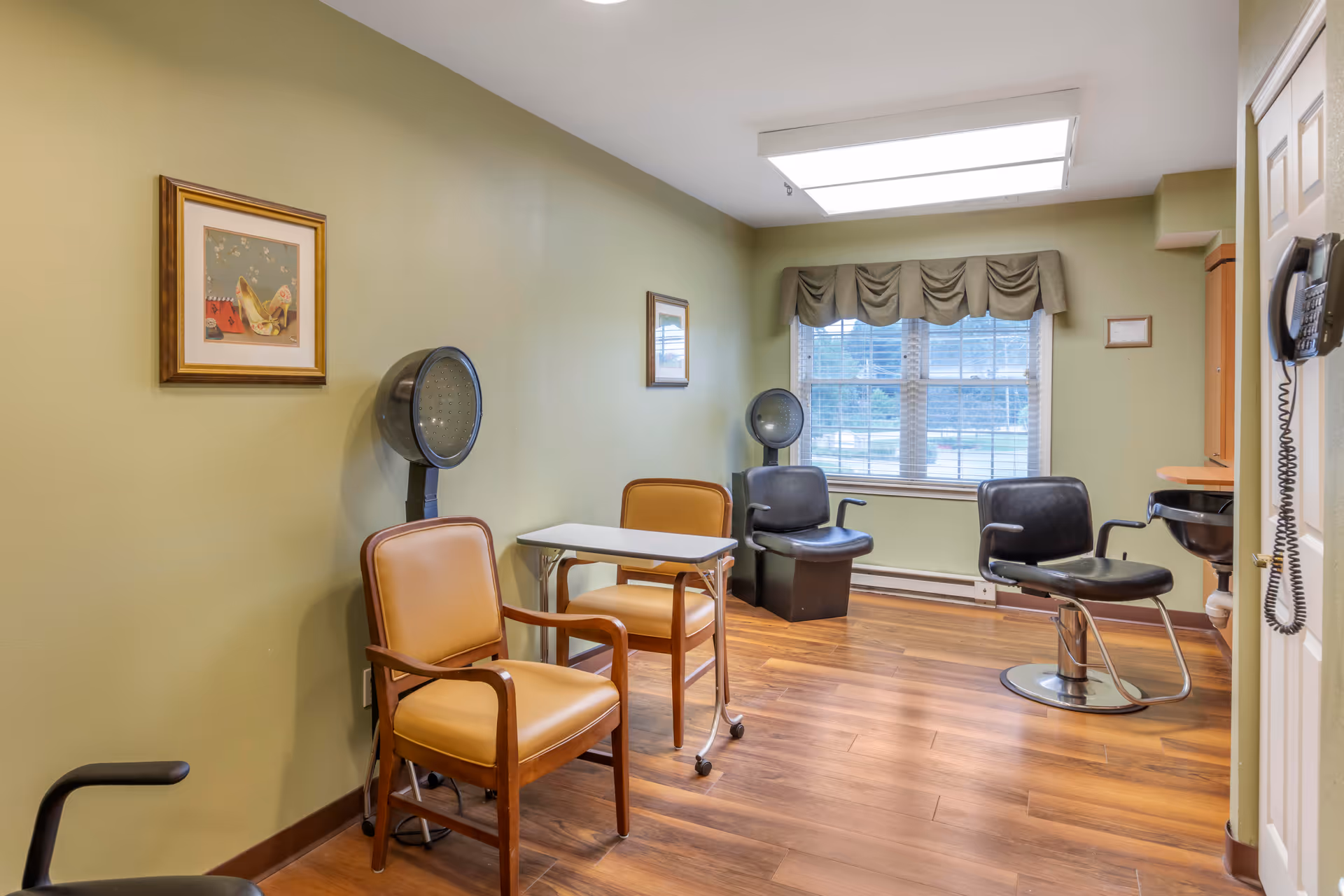 A small salon room with light green walls and wood flooring. The room contains two vintage hair dryer chairs with hooded dryers, two tan chairs with wooden arms, a small white rolling table, a black salon chair with a footrest, a wall-mounted phone, and a window with a valance letting in natural light. There are framed pictures on the walls.