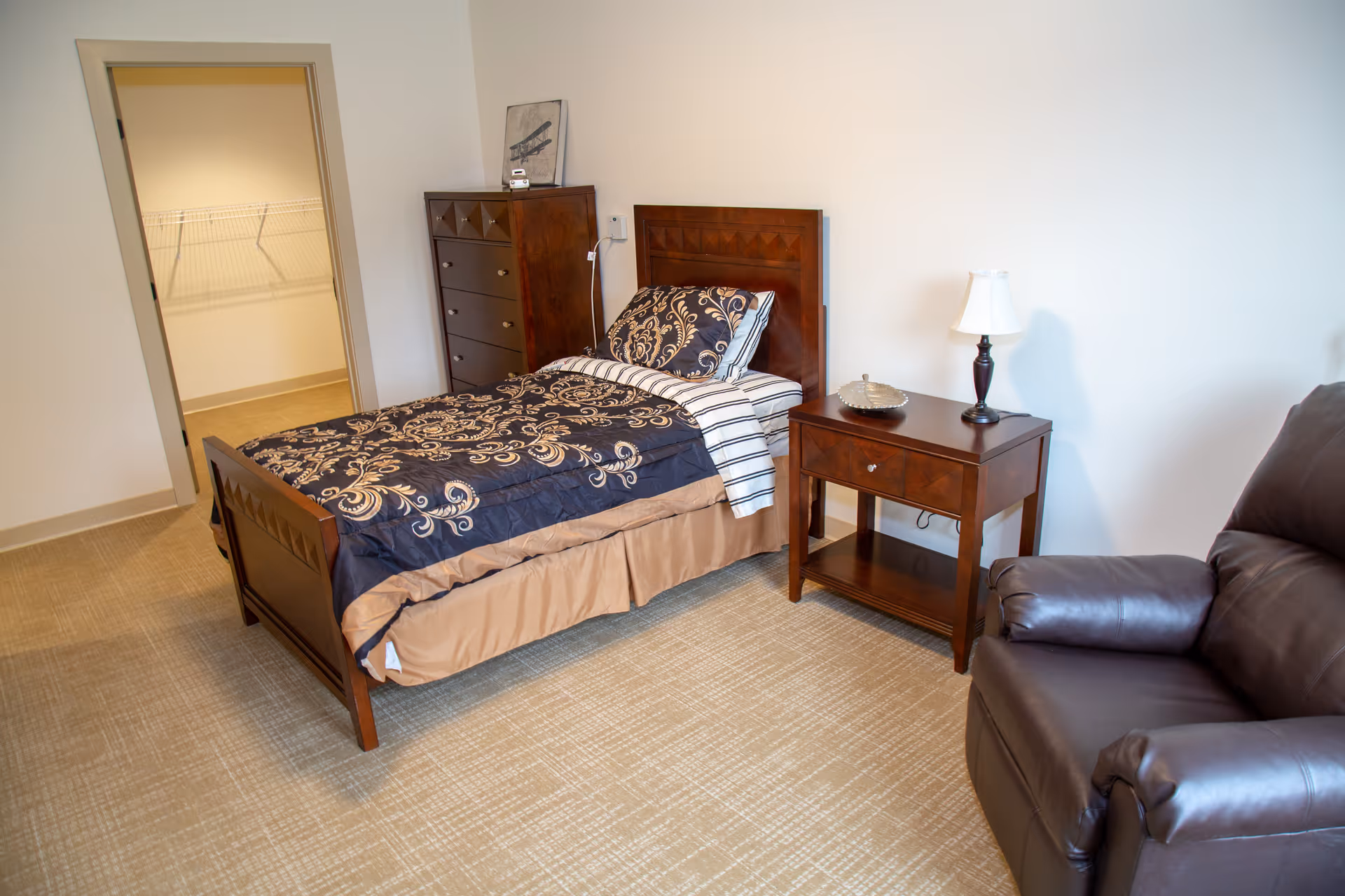 A bedroom with a single wooden bed featuring a dark blue and beige patterned comforter and striped pillow. Next to the bed is a wooden nightstand with a lamp and a decorative dish. There is a tall wooden dresser against the wall with a small framed picture on top. A brown leather recliner chair is positioned near the nightstand. The room has beige carpet and white walls, with an open doorway leading to a walk-in closet with wire shelving.
