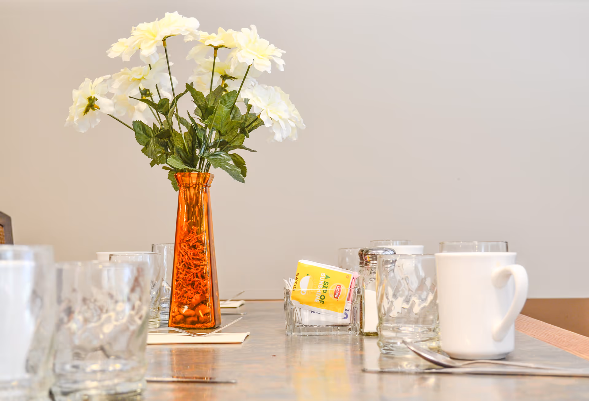 A dining table set with clear glasses, a white coffee mug, silverware, and a small glass container holding sugar packets. In the center of the table is a tall orange vase with white flowers. The background is a plain light-colored wall.