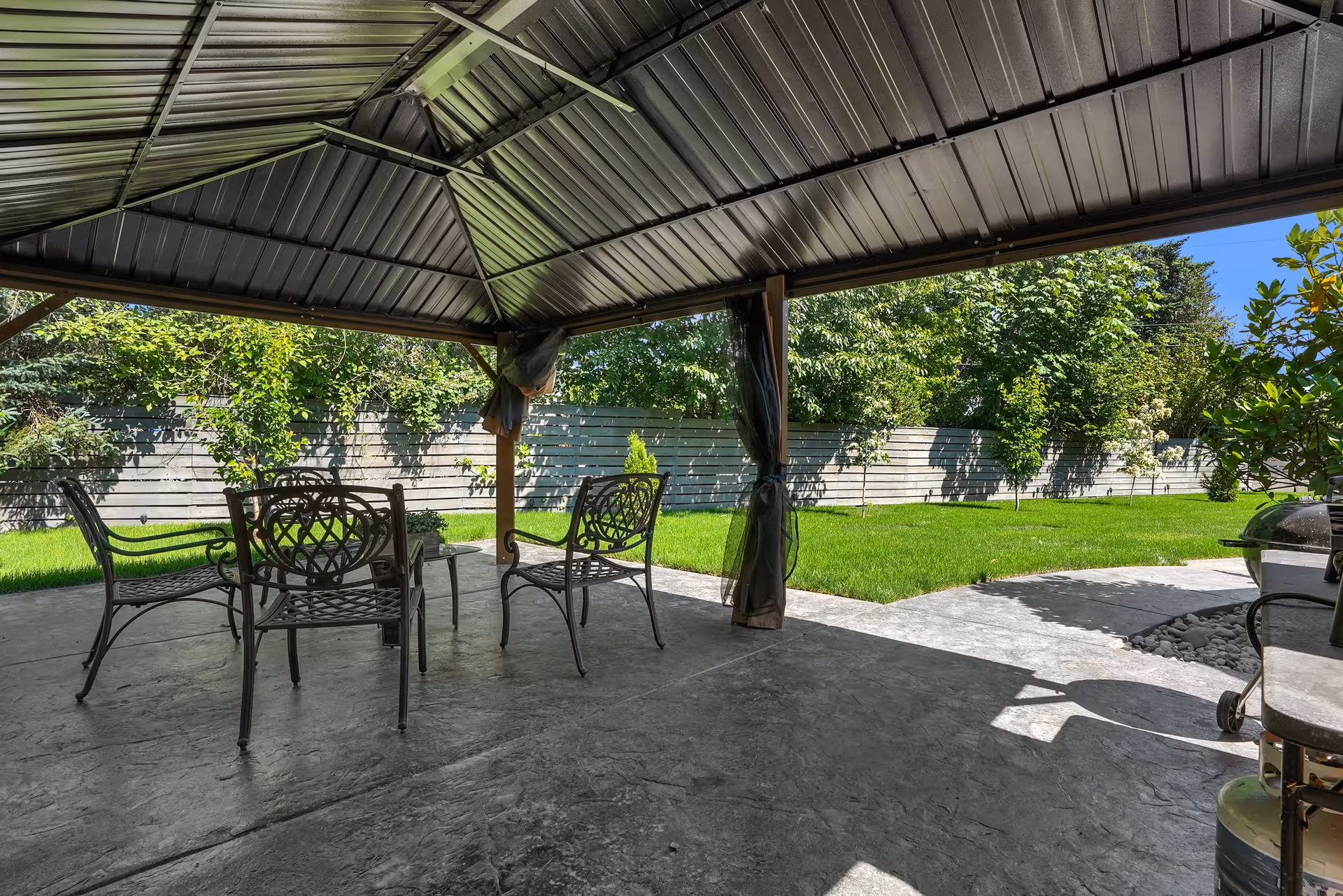 Outdoor covered patio area with metal roof and four metal chairs arranged around a small table. The patio overlooks a well-maintained grassy yard with trees and a wooden fence in the background under a clear blue sky.