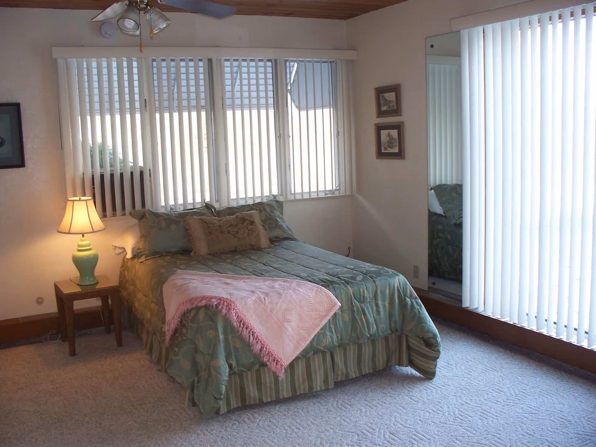 A bedroom with a bed covered in green patterned bedding and a pink throw blanket. There is a wooden nightstand with a green lamp on the left side of the bed. The room has large windows with vertical blinds and a ceiling fan with lights. Two framed pictures hang on the wall next to a large mirror.