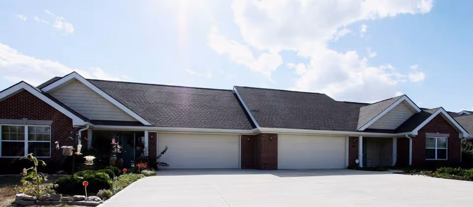 Exterior view of a single-story senior living facility building with brick walls, multiple garage doors, and a well-maintained driveway under a partly cloudy sky.