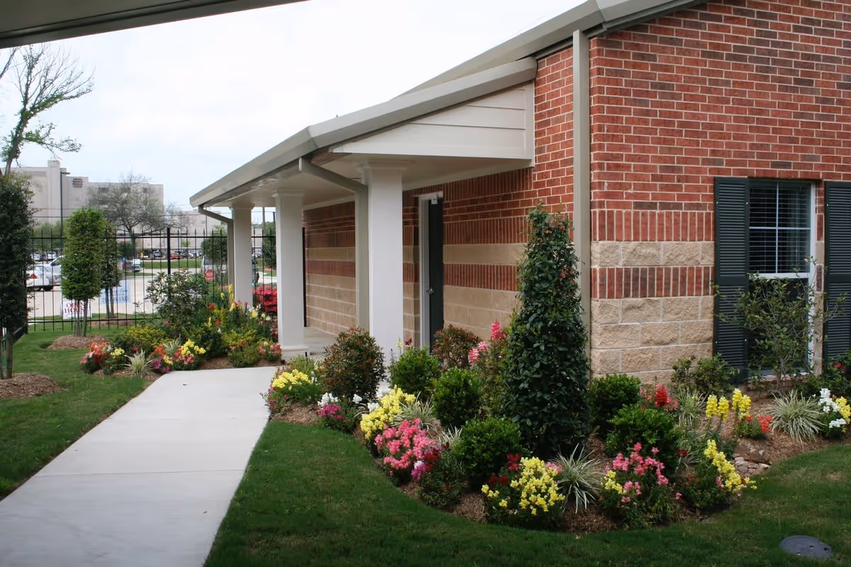 Exterior view of a brick building with a covered walkway supported by white columns. The walkway is bordered by well-maintained flower beds with colorful flowers and green shrubs. There is a window with dark green shutters on the building, and a black metal fence is visible in the background along with some trees and parked cars.