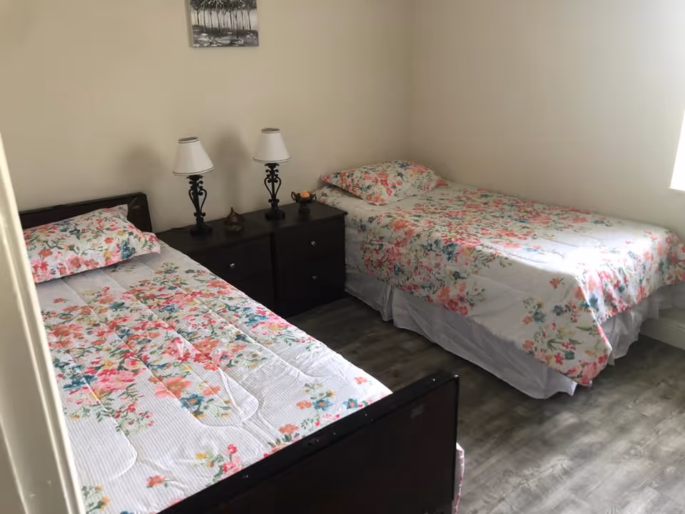 A small bedroom with two single beds placed parallel to each other, each covered with floral patterned bedspreads and matching pillows. Between the beds is a dark wooden nightstand with two decorative table lamps and a small decorative item. The room has light-colored walls, a wooden floor, and a small framed picture hanging above the nightstand.
