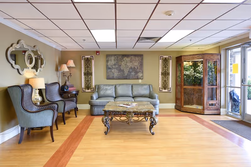 A bright lobby-style sitting area with a sofa, two armchairs, an ornate coffee table, wall art, and a glass entry door.