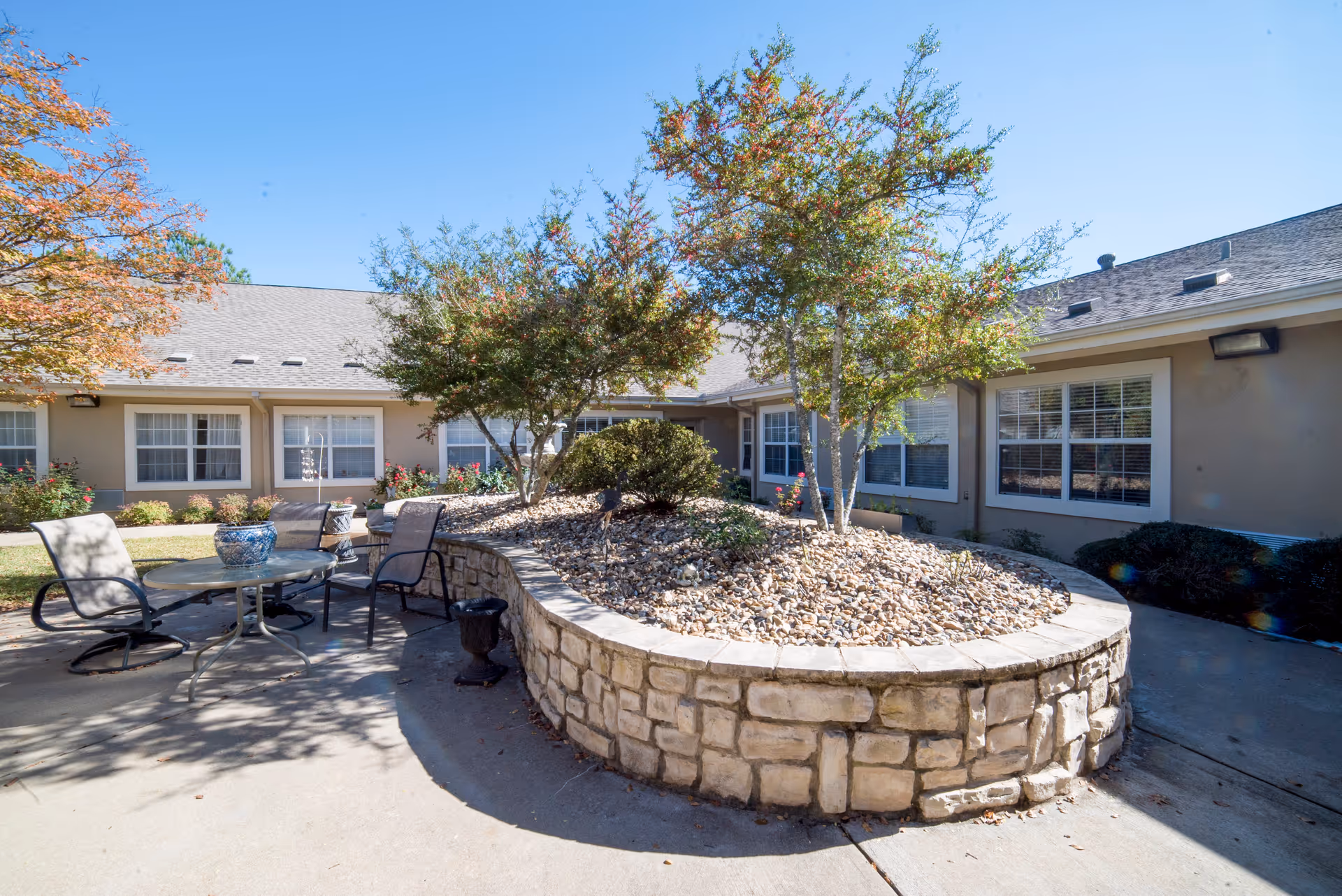 Sunny courtyard with a raised stone planter, small trees, and a patio table and chairs in front of a one-story senior living building.